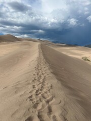 Footprints lead across towering sand dunes under a dramatic sky, capturing the solitude and raw beauty of a desert landscape just before a storm. Perfect for travel, nature, and adventure themes.