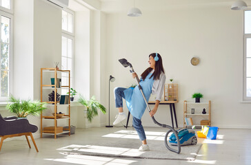 Young funny smiling woman housewife with earphones cleaning home with vacuum cleaner having fun,...