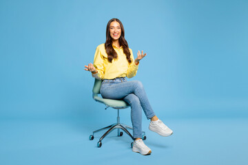 Positive young European woman talking and gesturing, looking at camera while sitting on chair over...