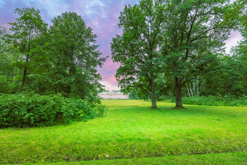 A serene evening view of green trees, grass, and a white palace under the setting sun. The sky is painted with soft pink and purple, creating a peaceful scene