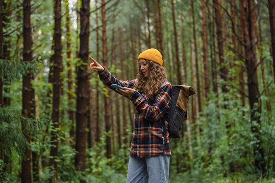 Woman wearing yellow beanie and plaid shirt uses her phone to navigate while pointing in the forest, blending technology with outdoor exploration.
