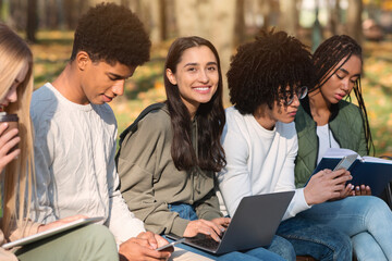 Multiracial teen friends spending time outdoors, using gadgets and reading books at park