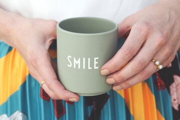 A detail of hands holding a sage green mug with "SMILE" printed on it. The person wears a pearl ring and a colorful, pleated skirt. The image exudes warmth and positivity.