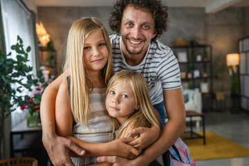 Fototapeta premium Portrait of daughters and dad stand and hug with backpacks at home