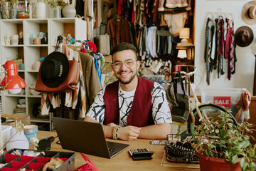 Man seated at desk in small boutique, smiling while surrounded by various clothing items and accessories, creating a cozy, welcoming atmosphere in the workspace with plants and colorful decor