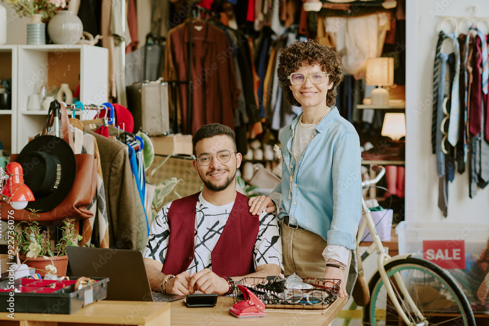 Wall mural Two smiling people posing in diverse and eclectic vintage clothing store surrounded by various clothes and accessories adding lively mood