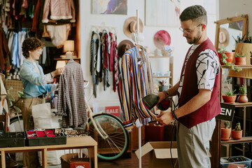 Focused man using handheld steamer on garment while woman works in background arranging items in creative shop setting filled with various merchandise and decorations