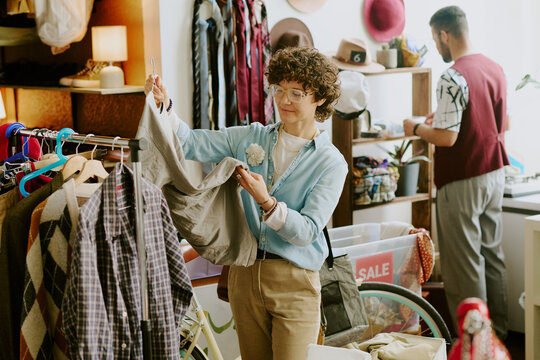 Man with curly hair examining clothing item in vibrant vintage store while another person browses items in background. Environments filled with diverse articles and sale signs