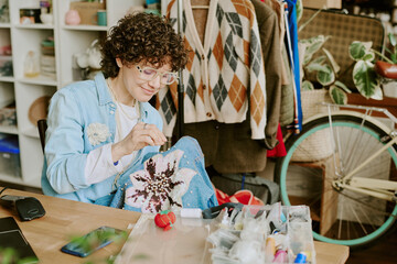 Woman wearing glasses, working on detailed floral graphic embroidery at wooden workspace surrounded...