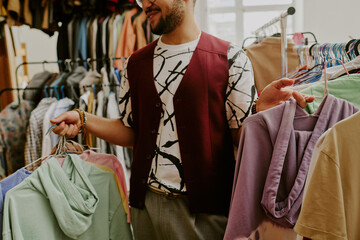 Smiling man browsing clothes in vibrant thrift store, holding hangers with various colorful garments while engaged in shopping experience, surrounded by racks of diverse fashion items