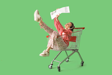 Displeased young fashionable woman in headphones with shopping cart reading newspaper on green...
