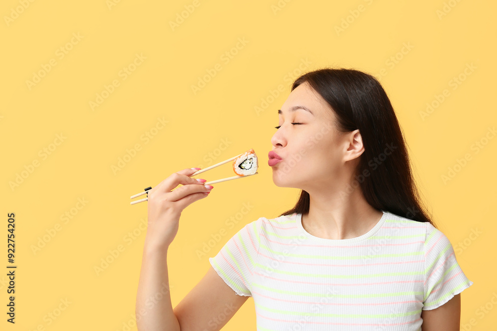 Poster beautiful young asian woman holding tasty sushi roll with chopsticks on yellow background