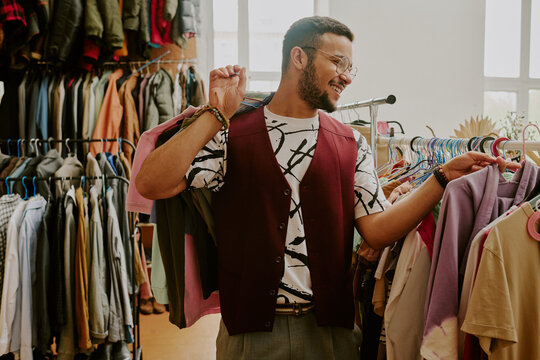 Smiling man wearing glasses shopping in a clothing store, surrounded by various garments on hangers, holding a jacket over his shoulder and enjoying the experience