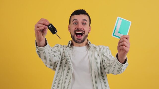 Young Caucasian lifting the new vehicle driver license L plate and a car key, with a big smile, being happy and proud of passing the driving school exam, looking at camera on a yellow background.