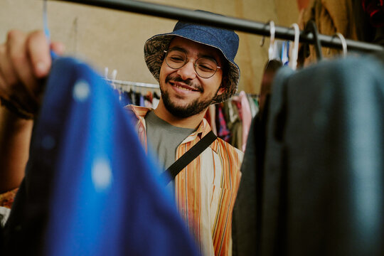 Young man wearing glasses with casual clothing and hat browsing clothes in thrift store while smiling cheerfully, engaged in the joy of shopping for unique apparel