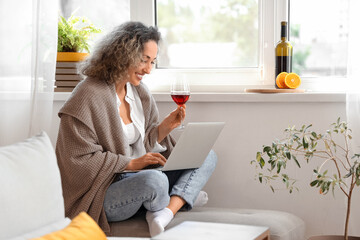 Beautiful middle-aged woman with glass of wine using laptop at home