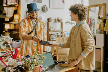Smiling couple at checkout counter in cozy boutique using card payment machine, one wearing bucket hat and colorful clothes, the other with glasses and short hair, exchanging friendly conversation