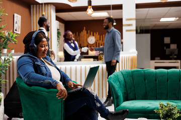 Smiling happy African American woman traveler resting in hotel lobby using headphones and laptop, wathing movie online or talking with family via video call. Tourist using free wifi hotspot at resort