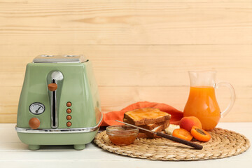 Green toaster with bread slices, apricots, jam and jug of juice on white wooden table