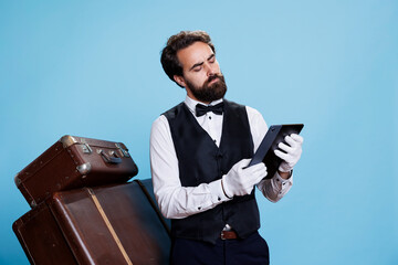 Classy porter reads guest list on device, acting like a professional and providing excellent concierge services. Bellboy with formal attire exudes sense of reliability, using tablet on camera.