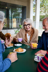 Vertical. Mature grey hair woman smiling enjoying coffee with group friends on terrace of old people home. Meeting of senior Caucasian group persons sitting together having snack or breakfast outdoor