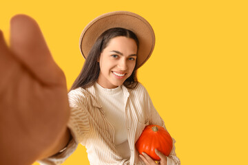 Stylish young woman with fresh pumpkin taking selfie on yellow background