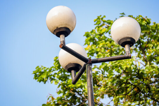 A close-up of a modern street lamp with three round bulbs, set against a clear blue sky and green foliage. The slightly rusted metal adds a touch of urban charm