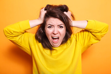 Close-up of desperate woman screaming and pulling her hair with her hands. Angry caucasian lady with a displeased and dramatic expression on her face, standing over isolated orange background.