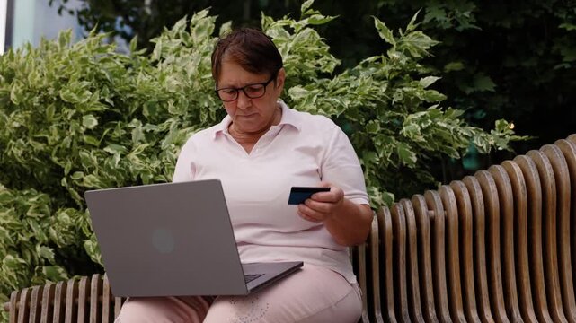 An older woman wearing glasses and a pink polo shirt is sitting on a modern wooden bench outdoors. She is using a laptop and holding a credit card, making an online purchase.