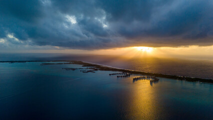 Low clouds loom over tropical island resorts featuring overwater villas and bungalows. The sunrise can be seen over the ocean behind them. 