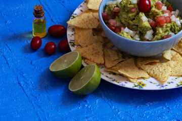Meal consisting of guacamole, nachos, cherry tomatoes, lime and olive oil served on a textured blue table