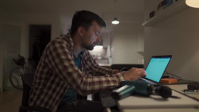 Cervical neck ache, nerve arthritis. Man studying late through night at laptop computer with cervical pain. Exhausted guy relaxing his neck using gun massager while overworking at notebook computer. 