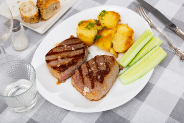 Deep-fried beef chop tenderloin is on round plate. Meat garnished with baked roasted potatoes, fresh cucumber slices and decorated with edible green, served with red wine