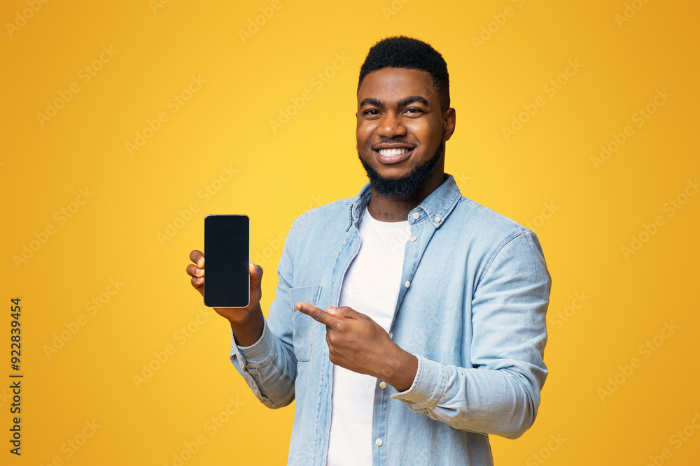 Poster handsome african american guy demonstrating smartphone with black screen and pointing on it, yellow 