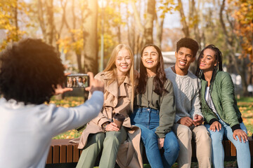 Black guy teenager taking photo of his happy international friends in autumn public park