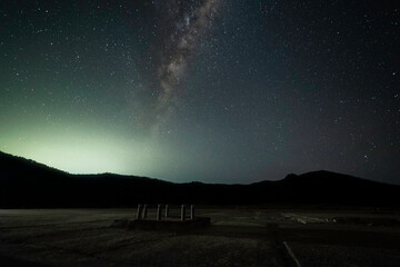 The Milky Way galaxy above a silhouette of mountains with part of a ruined tracking station in the foreground. You can see a glow of light over the mountains in the distance with a starry sky above.