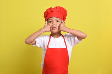 Cute little chef showing thumbs up isolated on yellow background. Asian boy wearing red apron and hat. Great little chef