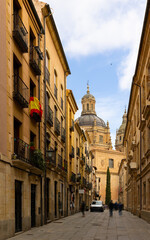 Obraz premium Picturesque cityscape of historic area of Salamanca overlooking people walking along typical narrow paved street leading to baroque building of La Clerecia with huge dome on sunny spring day, Spain.