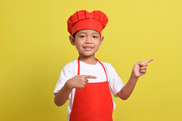 Cute little chef showing Blank Space Next to with pointing hand gesture isolated on yellow background. Asian boy wearing red apron and hat. Offer concept. Advertising banner