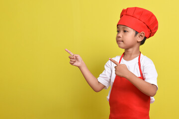 Cute little chef showing Blank Space Next to with pointing hand gesture isolated on yellow background. Asian boy wearing red apron and hat. Offer concept. Advertising banner