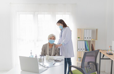 Female doctor in white coat comforting elderly patient in bright medical office. Both wearing face masks. doctor use stethoscope listening lung of patient, and pandemic safety measures in hospital.