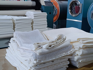 Folded white clothes, sheets, tablecloths, in an industrial laundry with machinery behind.