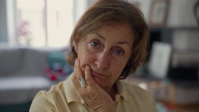 Older woman with a serious expression resting her face on her hand, indoors, close-up of her thoughtful look, wearing casual yellow polo shirt, home environment, soft lighting