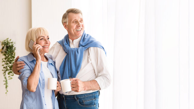 Mature woman talking on phone, embracing husband and drinking tea near window, free space