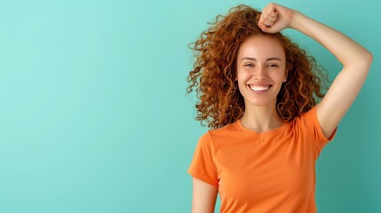 Fototapeta premium Smiling woman with curly hair in orange t-shirt, raising her arm against a teal background, expressing happiness and triumph.