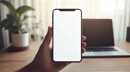 Obraz premium Hand Holding Smartphone with Blank Screen Mockup, Blurred Laptop and Plant Background.