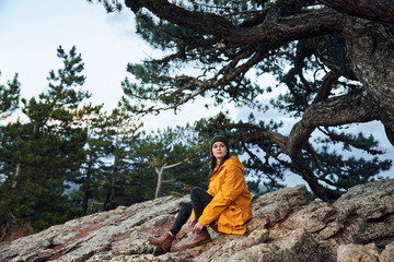 A woman in a stylish yellow coat enjoying nature sitting on a rock in a peaceful pine forest landscape