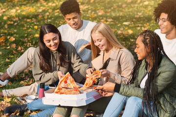 Group of positive young international friends eating take away pizza at public park