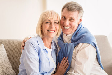 Happy mature couple embracing and smiling to camera, sitting on sofa at home