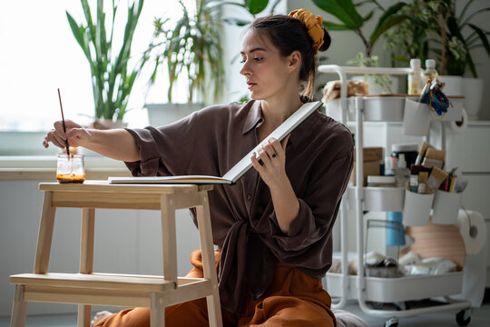 Inspired woman artist painting with watercolor paint in sketchbook, learns new technique in drawing workshop. Creative girl illustrator painter sits on floor working in art studio. Handmade hobby, DIY